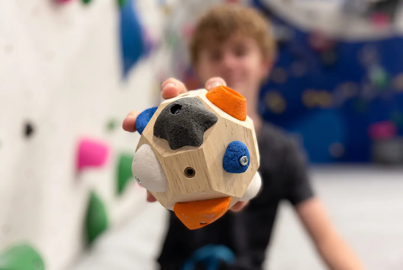 A young climber holding the Boulder Block up to the camera, the multi-faceted wooden training block with its colorful holds in sharp focus while the bouldering gym blurs behind them.