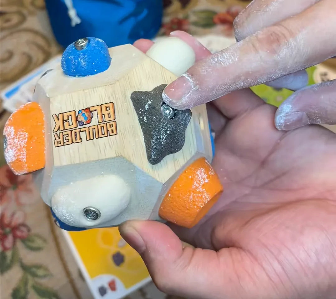 A chalky hand holding the Boulder Block, route cards from the Challenge Deck on the rug beside it
